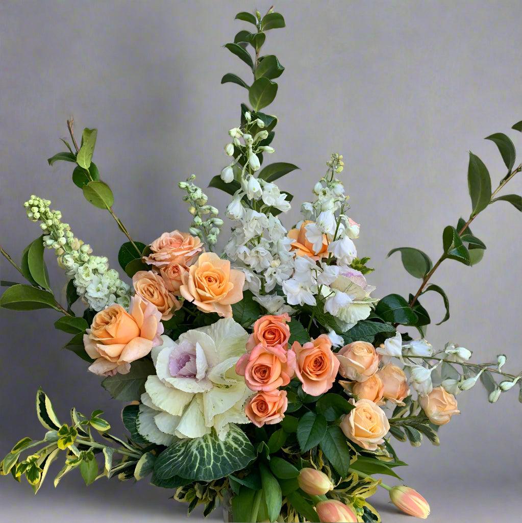 Elegant floral arrangement titled 'Peachy Love' featuring peach roses, white blooms, and lush greenery. Perfect for gift giving and celebrations.