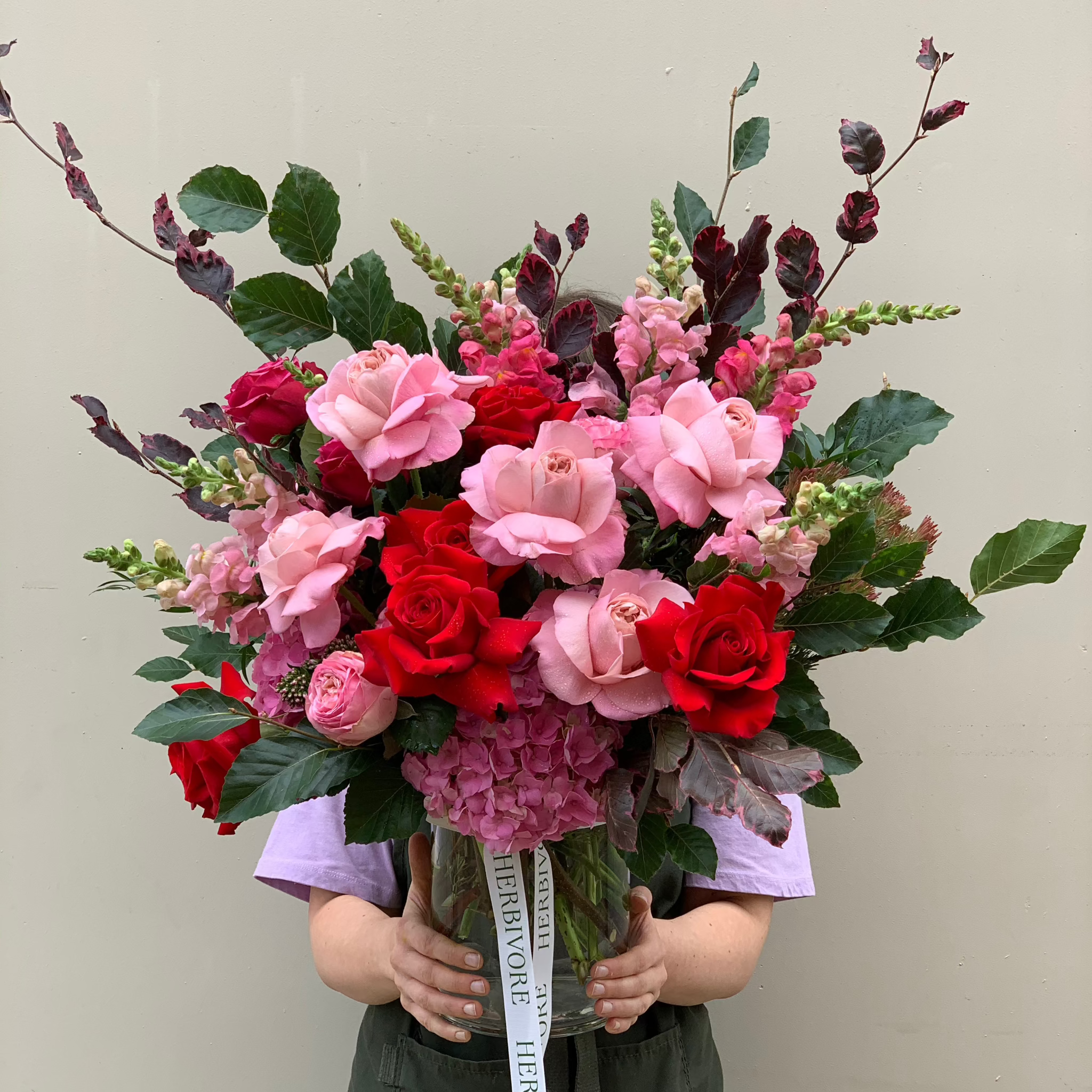 Herbilicious fresh floral arrangement featuring pink and red roses, snapdragons, hydrangea, and lush summer foliage, perfect for celebrations.