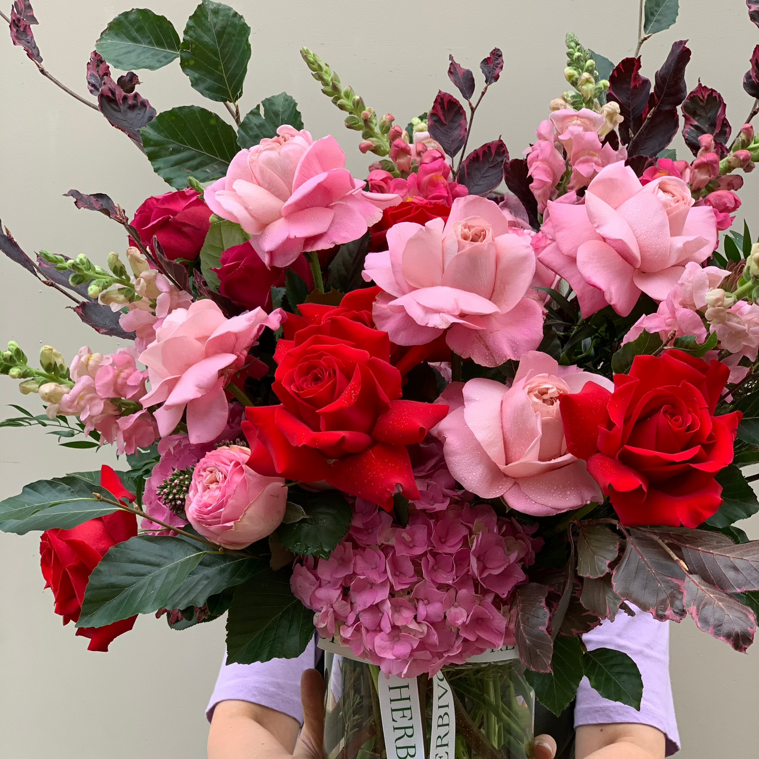 Herbilicious fresh floral arrangement featuring pink and red roses, snapdragons, hydrangeas, and seasonal summer foliage in a clear vase.