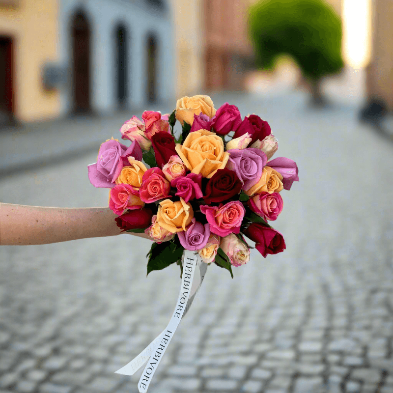 Rose Disco bouquet featuring a vibrant mix of colorful roses, including pink, orange, red, and yellow, held on a cobblestone street backdrop.