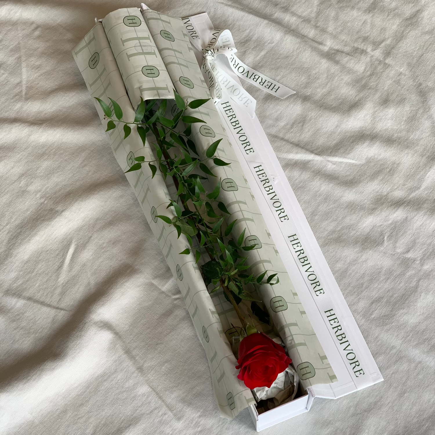 Single red rose elegantly presented in a white box with decorative greenery and branded wrapping, ideal for gift giving.